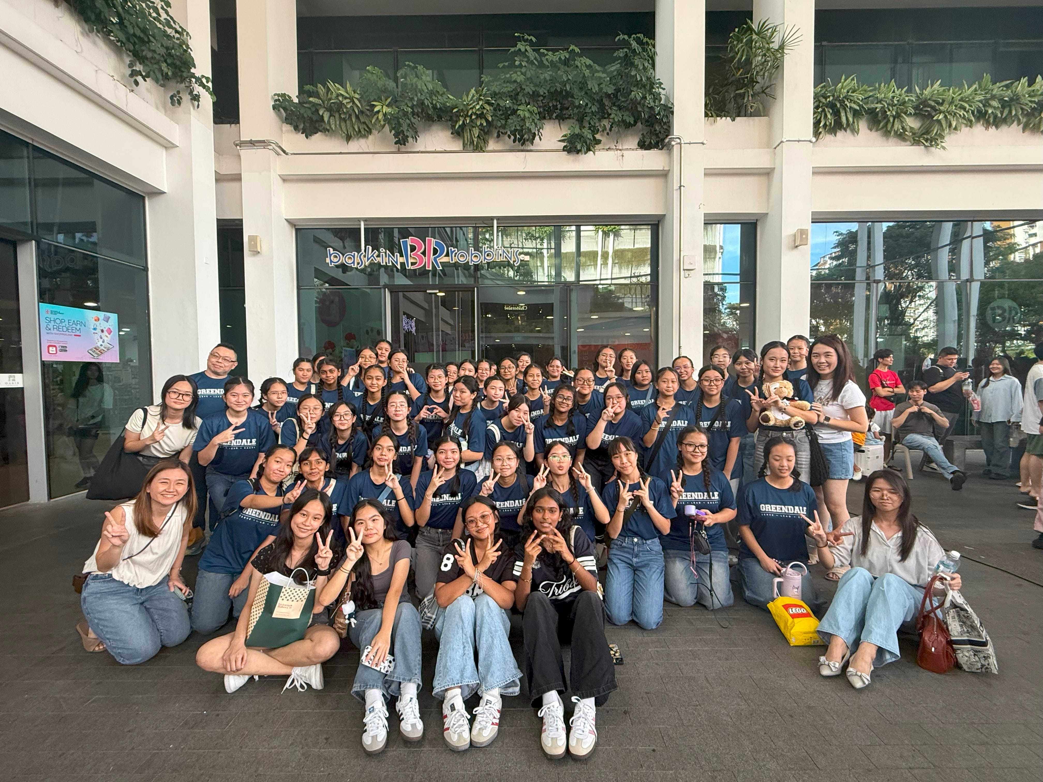 Large group of young people in matching navy Greendale shirts posing with peace signs outside a Baskin-Robbins store.